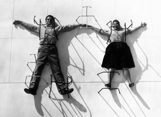 Charles e Ray Eames sdraiati a terra, tenendosi per mano, e intrappolati dalle strutture delle loro sedie